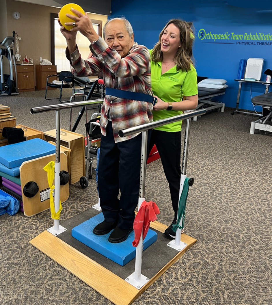 Picture of older gentleman standing on a balancing device between stability bars, and holding a ball overhead while his OTR therapist stands behind.